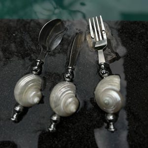 May include: A set of three stainless steel utensils: a spoon, knife, and fork. Each utensil features a decorative handle made from a polished, pearlescent seashell. The silverware is arranged on a dark, wet surface.