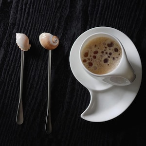 Puede incluir: Una taza de café de cerámica blanca con un asa y platillo únicos, llena de una bebida marrón espumosa. Dos cucharas decorativas con conchas marinas están a la izquierda. El fondo es una superficie oscura y texturizada.