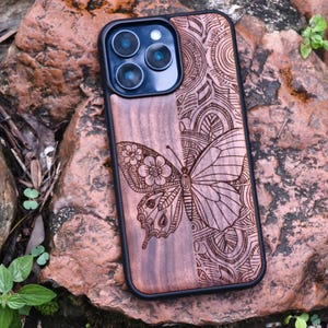 May include: A wooden phone case with a butterfly design. The case is split into two sections, one with a butterfly and floral design, and the other with an intricate pattern. The case has a black border and a camera cutout.