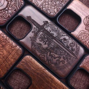 May include: Wooden phone cases with intricate engraved designs. One case features a sword and shield emblem with a detailed illustration. Other cases showcase various patterns and symbols. The cases are set in a black frame.
