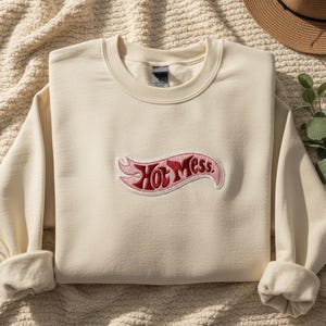 May include: Cream-coloured sweatshirt featuring the embroidered phrase "Hot Mess" in a red and pink banner design. The long-sleeved top has a crew neck and rolled cuffs. A straw hat and greenery are in the background.