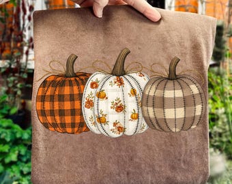 May include: A brown pillow featuring three decorative pumpkins. The pumpkins are in orange plaid, white floral, and brown plaid patterns. Each pumpkin has a brown stem and is tied with twine. The pillow is held up by a hand.
