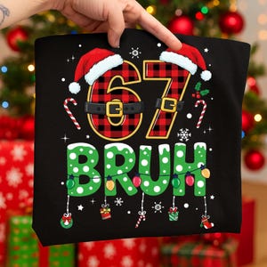 May include: Black t-shirt with a festive Christmas design. The number "67" is in red and black plaid, topped with Santa hats. The word "BRUH" is in green with white polka dots, decorated with Christmas lights and ornaments.