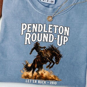 May include: A blue jean Comfort Colors t-shirt featuring the text "PENDLETON ROUND-UP" and a graphic of a cowboy on a bucking horse. The text "LET'ER BUCK - 1910" is below the illustration. A gold necklace is draped across the shirt.