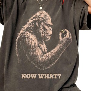 May include: A charcoal gray short-sleeve t-shirt featuring a graphic illustration of a hairy primate looking at a small ring held in its hand. The text "NOW WHAT?" is printed in a light beige color below the graphic. The shirt has a crew neckline.