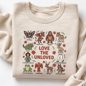 May include: A beige crewneck sweatshirt featuring a central graphic with the text LOVE THE UNLOVED surrounded by illustrations of cryptids including Mothman, Jersey Devil, Bigfoot, Chupacabra, Hodag, Jackalope, Flatwoods Monster, and others.