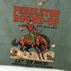 May include: A moss green t-shirt with a vintage graphic of a cowboy riding a horse, with the text "PENDLETON ROUND-UP" and "LET 'ER BUCK! AMERICA'S BIGGEST WESTERN SHOW". The shirt has a crew neck and short sleeves.