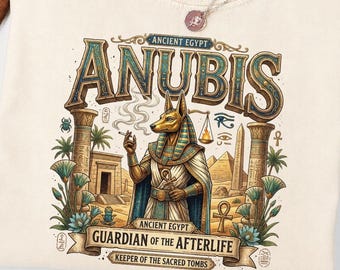 May include: A cream-colored t-shirt featuring a graphic of the Egyptian deity Anubis. The design includes the text ANUBIS, ANCIENT EGYPT, GUARDIAN OF THE AFTERLIFE, and KEEPER OF THE SACRED TOMBS, alongside illustrations of pyramids, columns, and plants.
