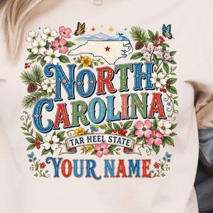 Puede incluir: Camiseta de color marfil con un dise&ntilde;o floral colorido y las palabras "North Carolina" en una fuente decorativa. El dise&ntilde;o incluye un mapa de Carolina del Norte, mariposas y el texto "Tar Heel State" y "Your Name".