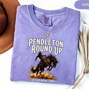 May include: A violet Comfort Colors t-shirt with the text "Pendleton Round-Up" and a graphic of a cowboy riding a bucking bronco. The text "Let'er Buck - 1910" is below the graphic. A brown cowboy hat and a gold necklace are also in the image.