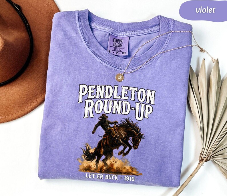 May include: A violet Comfort Colors t-shirt with the text "Pendleton Round-Up" and a graphic of a cowboy riding a bucking bronco. The text "Let'er Buck - 1910" is below the graphic. A brown cowboy hat and a gold necklace are also in the image.