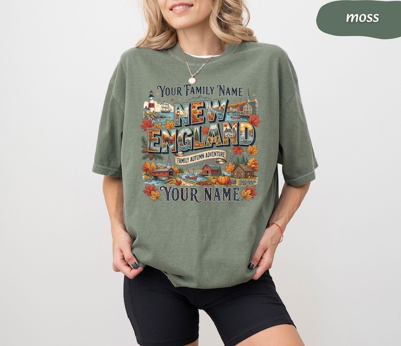 May include: A moss green short-sleeve t-shirt featuring a graphic print with the text "YOUR FAMILY NAME NEW ENGLAND FAMILY AUTUMN ADVENTURE YOUR NAME". The design includes illustrations of a lighthouse, autumn foliage, houses, and a pumpkin.