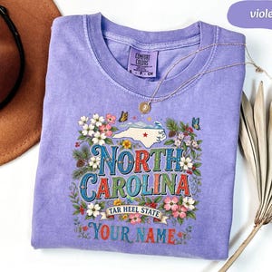 Puede incluir: Una camiseta violeta Comfort Colors con un dise&ntilde;o floral. El dise&ntilde;o incluye las palabras "North Carolina" y "Tar Heel State", un mapa de Carolina del Norte, flores y mariposas. Tambi&eacute;n se ven las palabras "Your Name".