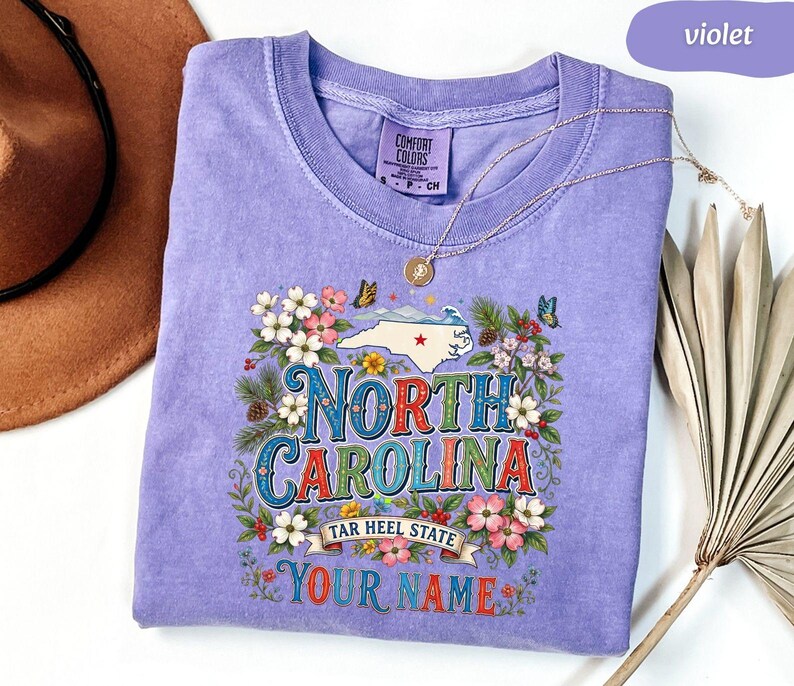 Puede incluir: Una camiseta violeta Comfort Colors con un dise&ntilde;o floral. El dise&ntilde;o incluye las palabras "North Carolina" y "Tar Heel State", un mapa de Carolina del Norte, flores y mariposas. Tambi&eacute;n se ven las palabras "Your Name".
