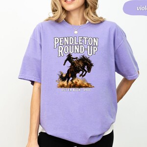 May include: A lilac-coloured t-shirt with the text "PENDLETON ROUND-UP" above a rodeo illustration of a cowboy on a bucking bronco. The text "LET ER BUCK - 1910" is below the illustration.