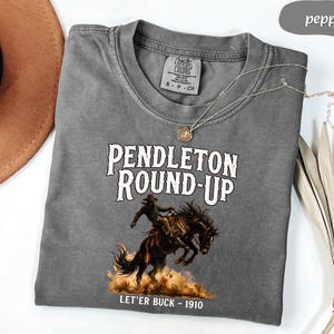 May include: A grey Comfort Colors t-shirt with the text "PENDLETON ROUND-UP" and an illustration of a cowboy on a bucking horse. The text "LET'ER BUCK - 1910" is below the illustration. A brown hat and a necklace are also visible.