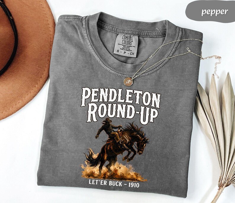 May include: A grey Comfort Colors t-shirt with the text "PENDLETON ROUND-UP" and an illustration of a cowboy on a bucking horse. The text "LET'ER BUCK - 1910" is below the illustration. A brown hat and a necklace are also visible.
