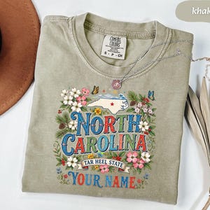 Puede incluir: Camiseta de color caqui con un dise&ntilde;o floral y las palabras "NORTH CAROLINA" en una fuente vintage colorida. El dise&ntilde;o incluye un mapa de Carolina del Norte, mariposas y el texto "TAR HEEL STATE" y "YOUR NAME". Un collar plateado est&aacute; drapeado sobre la camiseta.