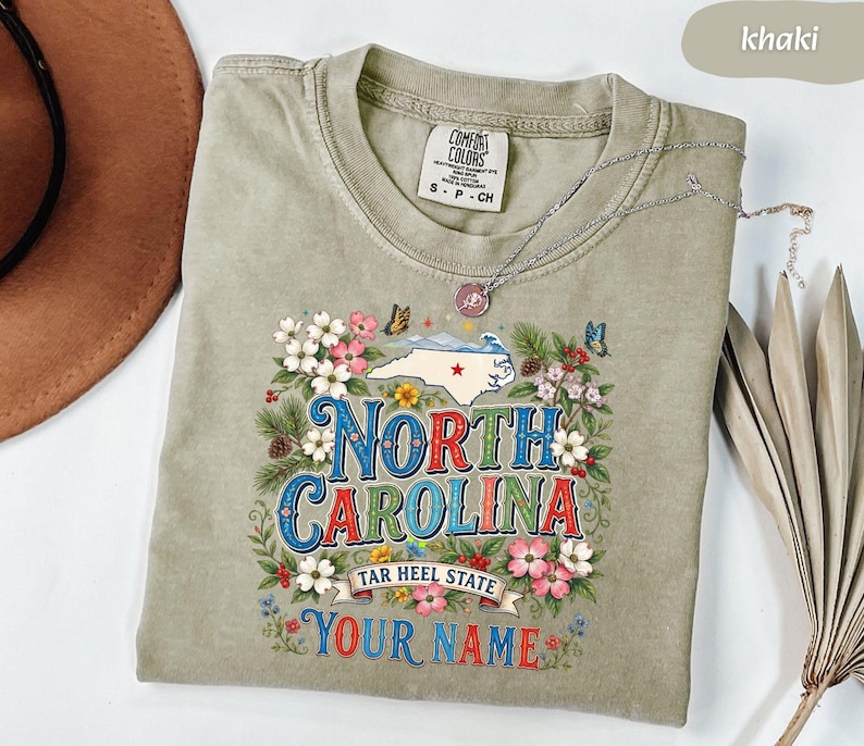 Puede incluir: Camiseta de color caqui con un dise&ntilde;o floral y las palabras "NORTH CAROLINA" en una fuente vintage colorida. El dise&ntilde;o incluye un mapa de Carolina del Norte, mariposas y el texto "TAR HEEL STATE" y "YOUR NAME". Un collar plateado est&aacute; drapeado sobre la camiseta.