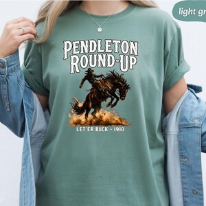 May include: Light green t-shirt with the text "Pendleton Round-Up" and an illustration of a cowboy on a bucking bronco. Below the image is the text "Let'er Buck - 1910". The shirt has short sleeves.