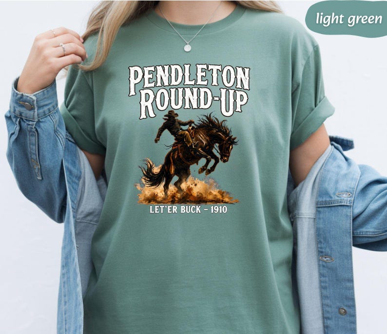 May include: Light green t-shirt with the text "Pendleton Round-Up" and an illustration of a cowboy on a bucking bronco. Below the image is the text "Let'er Buck - 1910". The shirt has short sleeves.