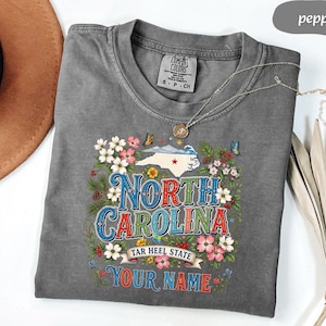 Puede incluir: Camiseta gris con un dise&ntilde;o floral que presenta el estado de Carolina del Norte. El dise&ntilde;o incluye el texto "North Carolina", "Tar Heel State" y espacio para un nombre personalizado. La camiseta tiene cuello redondo y mangas cortas.