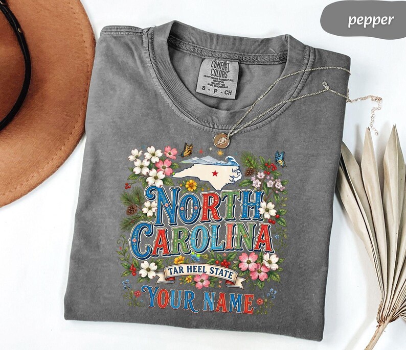 Puede incluir: Camiseta gris con un dise&ntilde;o floral que presenta el estado de Carolina del Norte. El dise&ntilde;o incluye el texto "North Carolina", "Tar Heel State" y espacio para un nombre personalizado. La camiseta tiene cuello redondo y mangas cortas.