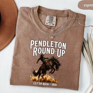 May include: Espresso-coloured t-shirt with the text "PENDLETON ROUND-UP" and a cowboy riding a bucking bronco illustration. The shirt also features the text "LET'ER BUCK - 1910". It is a Comfort Colors brand.