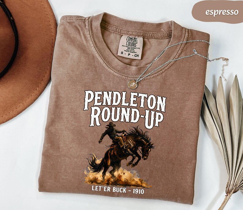May include: Espresso-coloured t-shirt with the text "PENDLETON ROUND-UP" and a cowboy riding a bucking bronco illustration. The shirt also features the text "LET'ER BUCK - 1910". It is a Comfort Colors brand.