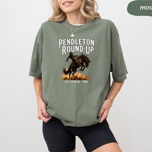 May include: A moss green t-shirt with the words "Pendleton Round-Up" and an illustration of a rodeo rider on a bucking bronco. The text "Let 'er Buck - 1910" is below the image. The shirt has a loose fit.