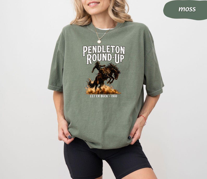 May include: A moss green t-shirt with the words "Pendleton Round-Up" and an illustration of a rodeo rider on a bucking bronco. The text "Let 'er Buck - 1910" is below the image. The shirt has a loose fit.