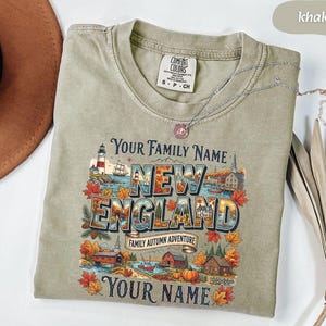 May include: A khaki-coloured crew neck t-shirt featuring a graphic print with the text "YOUR FAMILY NAME", "NEW ENGLAND", "FAMILY AUTUMN ADVENTURE", and "YOUR NAME". The design includes a lighthouse, autumn leaves, pumpkins, and houses.