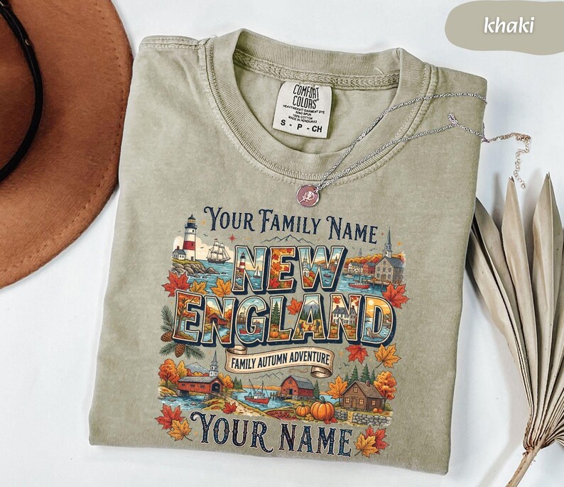 May include: A khaki-coloured crew neck t-shirt featuring a graphic print with the text "YOUR FAMILY NAME", "NEW ENGLAND", "FAMILY AUTUMN ADVENTURE", and "YOUR NAME". The design includes a lighthouse, autumn leaves, pumpkins, and houses.