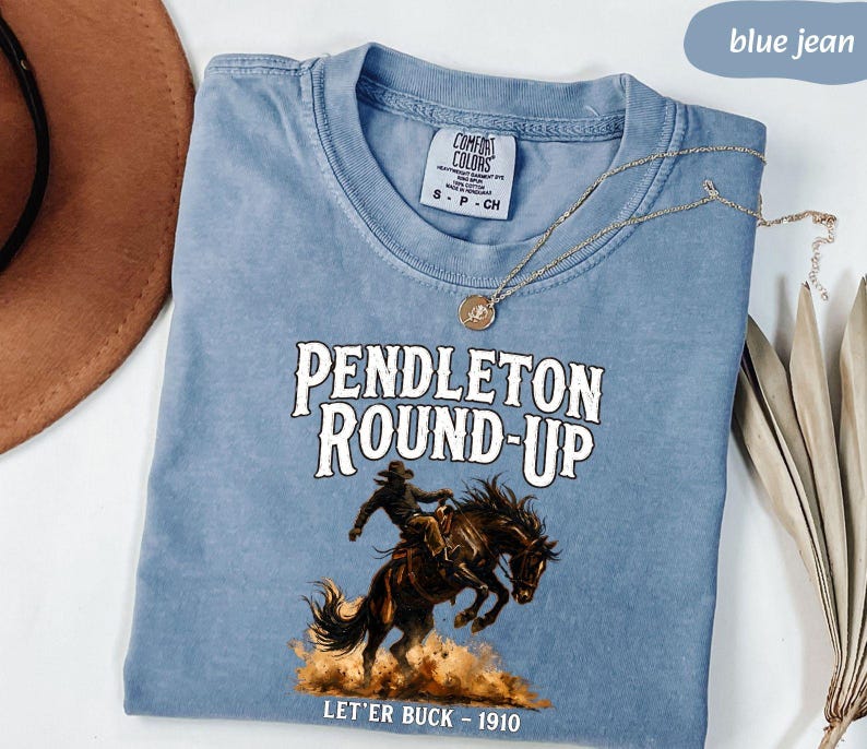 May include: A blue jean Comfort Colors t-shirt featuring the text "PENDLETON ROUND-UP" and a graphic of a cowboy on a bucking horse. The text "LET'ER BUCK - 1910" is below the illustration. A gold necklace is draped across the shirt.