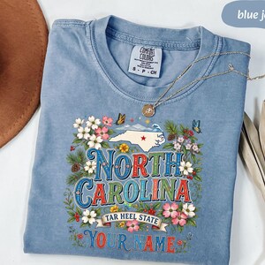 Puede incluir: Una camiseta azul vaquera Comfort Colors con un dise&ntilde;o floral y un mapa estatal. El dise&ntilde;o incluye las palabras "North Carolina" y "Tar Heel State", con espacio para un nombre. Un collar plateado descansa sobre la camisa.