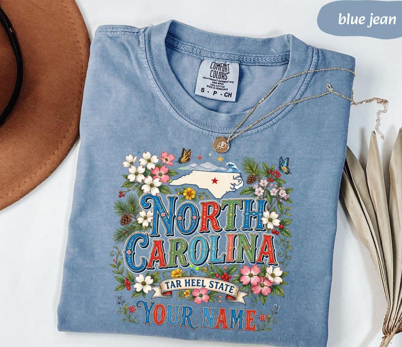 Puede incluir: Una camiseta azul vaquera Comfort Colors con un dise&ntilde;o floral y un mapa estatal. El dise&ntilde;o incluye las palabras "North Carolina" y "Tar Heel State", con espacio para un nombre. Un collar plateado descansa sobre la camisa.