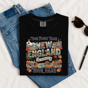 May include: A black t-shirt featuring a graphic with the text "YOUR FAMILY NAME", "NEW ENGLAND", "FAMILY AUTUMN ADVENTURE", and "YOUR NAME". The design includes illustrations of a lighthouse, a ship, autumn leaves, a pumpkin, and buildings.