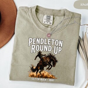 May include: Khaki t-shirt with the text "PENDLETON ROUND-UP" and an illustration of a cowboy on a bucking horse. The text "LET'ER BUCK - 1910" is below the image. A silver necklace with a pendant is draped across the shirt.