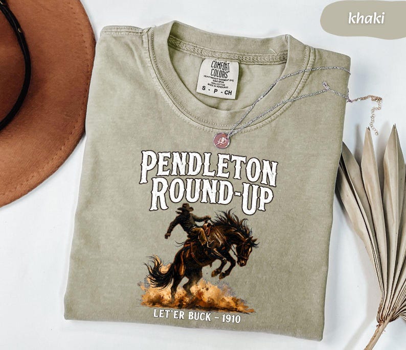 May include: Khaki t-shirt with the text "PENDLETON ROUND-UP" and an illustration of a cowboy on a bucking horse. The text "LET'ER BUCK - 1910" is below the image. A silver necklace with a pendant is draped across the shirt.