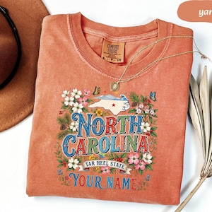 Puede incluir: Una camiseta color &ntilde;ame con un dise&ntilde;o floral alrededor de las palabras "NORTH CAROLINA" y un mapa del estado. La camiseta incluye el texto "TAR HEEL STATE" y espacio para un nombre. Un collar de oro est&aacute; drapeado sobre la camiseta.