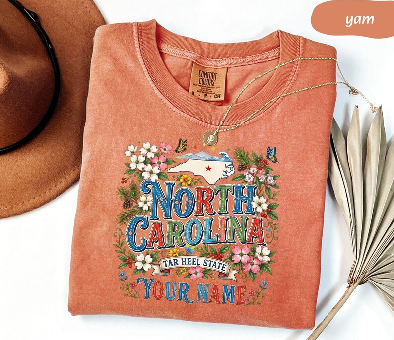 Puede incluir: Una camiseta color &ntilde;ame con un dise&ntilde;o floral alrededor de las palabras "NORTH CAROLINA" y un mapa del estado. La camiseta incluye el texto "TAR HEEL STATE" y espacio para un nombre. Un collar de oro est&aacute; drapeado sobre la camiseta.