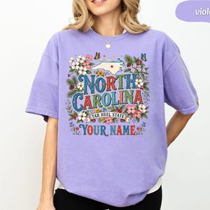 Puede incluir: Camiseta violeta con un dise&ntilde;o floral. El dise&ntilde;o incluye las palabras "North Carolina" y "Tar Heel State", un mapa de Carolina del Norte y espacio para un nombre. Mariposas y flores rodean el texto.