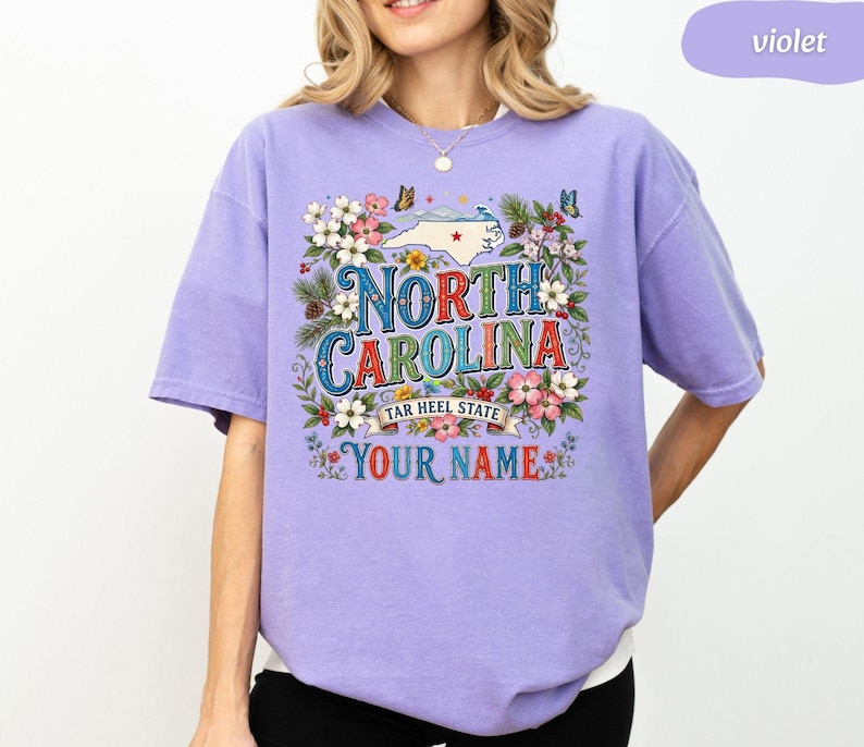 Puede incluir: Camiseta violeta con un dise&ntilde;o floral. El dise&ntilde;o incluye las palabras "North Carolina" y "Tar Heel State", un mapa de Carolina del Norte y espacio para un nombre. Mariposas y flores rodean el texto.