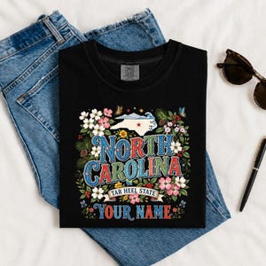 Puede incluir: Camiseta negra con un dise&ntilde;o floral colorido y las palabras "North Carolina" en una fuente vintage. El dise&ntilde;o incluye el contorno del estado, flores y el texto "Tar Heel State" y "Your Name".