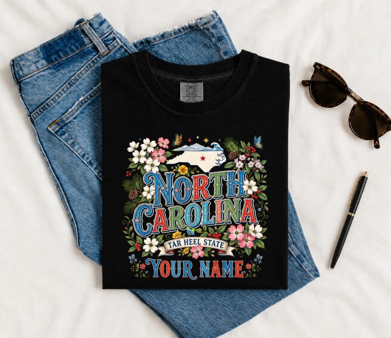 Puede incluir: Camiseta negra con un dise&ntilde;o floral colorido y las palabras "North Carolina" en una fuente vintage. El dise&ntilde;o incluye el contorno del estado, flores y el texto "Tar Heel State" y "Your Name".