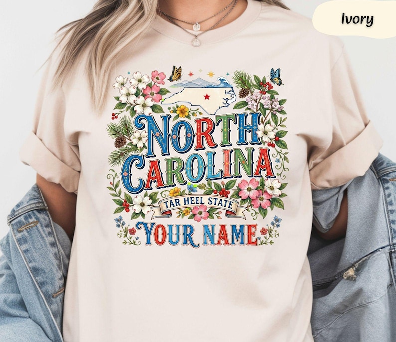 Puede incluir: Camiseta de color marfil con un dise&ntilde;o floral colorido y las palabras "North Carolina" en una fuente decorativa. El dise&ntilde;o incluye un mapa de Carolina del Norte, mariposas y el texto "Tar Heel State" y "Your Name".