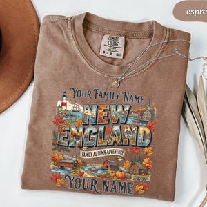 May include: An espresso-coloured short-sleeve t-shirt featuring a graphic print with the text "Your Family Name," "New England," "Family Autumn Adventure," and "Your Name." The design includes a lighthouse, autumn leaves, and houses.