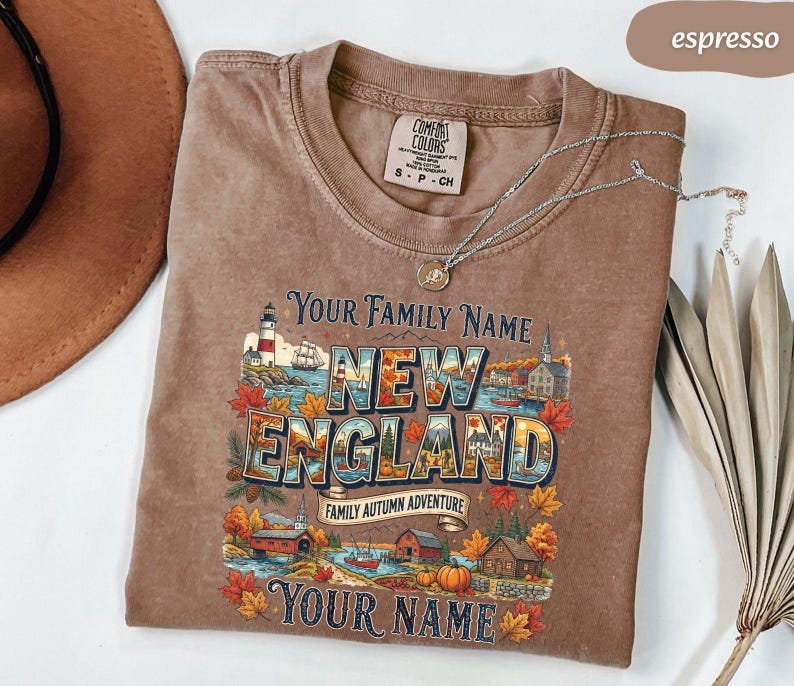 May include: An espresso-coloured short-sleeve t-shirt featuring a graphic print with the text "Your Family Name," "New England," "Family Autumn Adventure," and "Your Name." The design includes a lighthouse, autumn leaves, and houses.