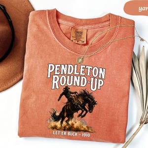 May include: A yam-coloured Comfort Colors t-shirt with the text "Pendleton Round-Up" above an illustration of a cowboy riding a bucking bronco. The text "Let'er Buck - 1910" is below the illustration. A gold necklace and a brown hat are also visible.