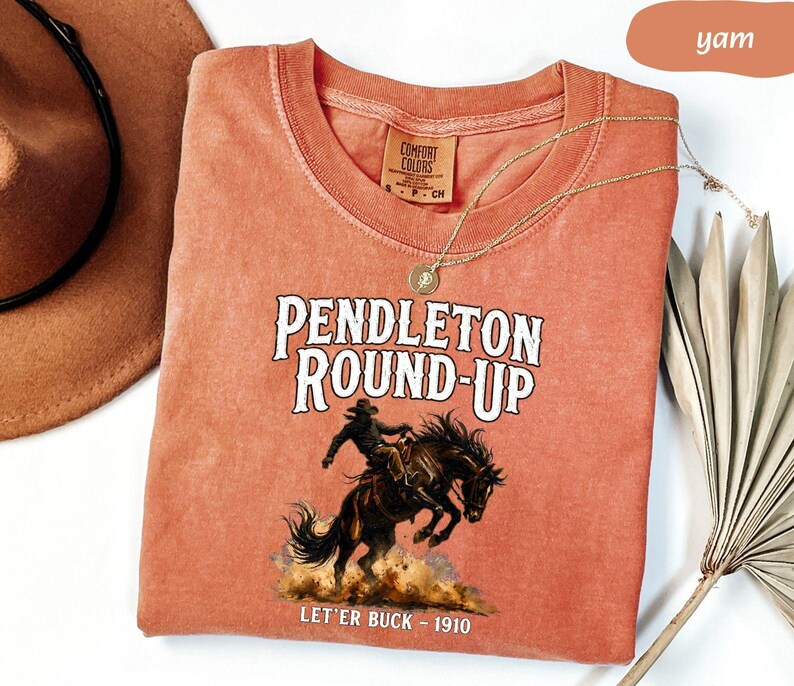 May include: A yam-coloured Comfort Colors t-shirt with the text "Pendleton Round-Up" above an illustration of a cowboy riding a bucking bronco. The text "Let'er Buck - 1910" is below the illustration. A gold necklace and a brown hat are also visible.